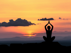 Woman sits on mountain with hands above head while looking at a sunrise.