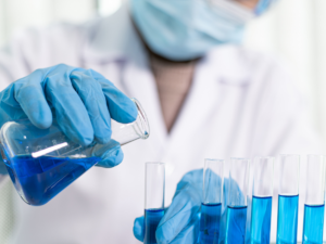 Women in a lab coat with a mask over math is pouring a blue substance in vials.