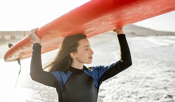 Portrait of a young woman in wetsuit carrying surboard above the head, preparing for surfing on the wild beach on a sunset near Sober Partners Drug and Alcohol Rehab