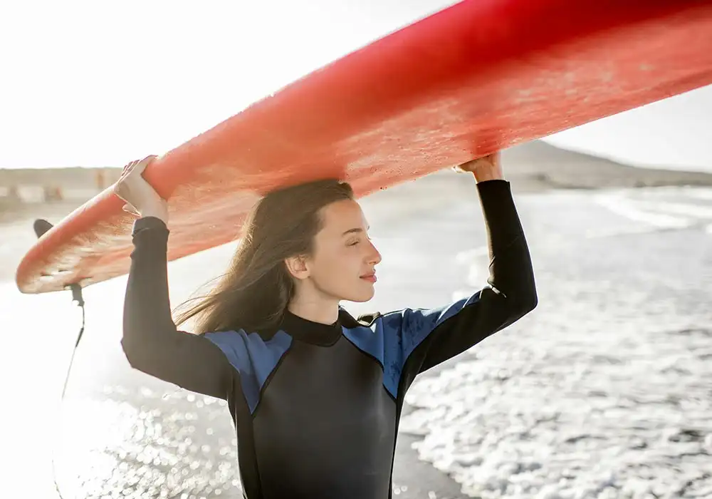 Portrait of a young woman in wetsuit carrying surboard above the head, preparing for surfing on the wild beach on a sunset near Sober Partners Drug and Alcohol Rehab